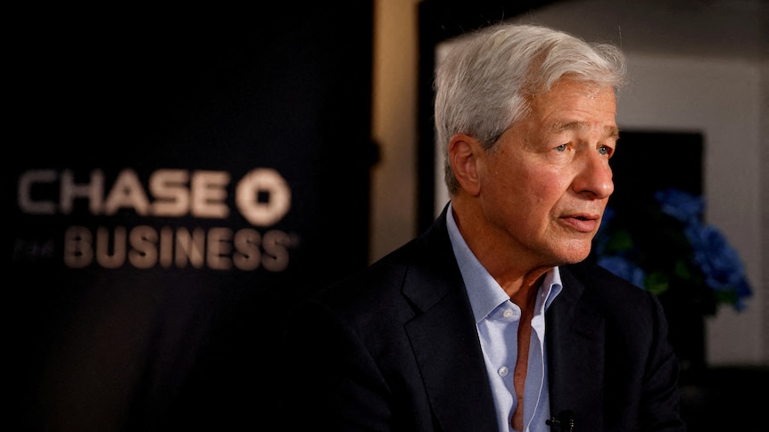 A picture of Jamie Dimon sitting in front of the JPMorgan Chase logo. He has white hair and black eyes. Wears a suit. 