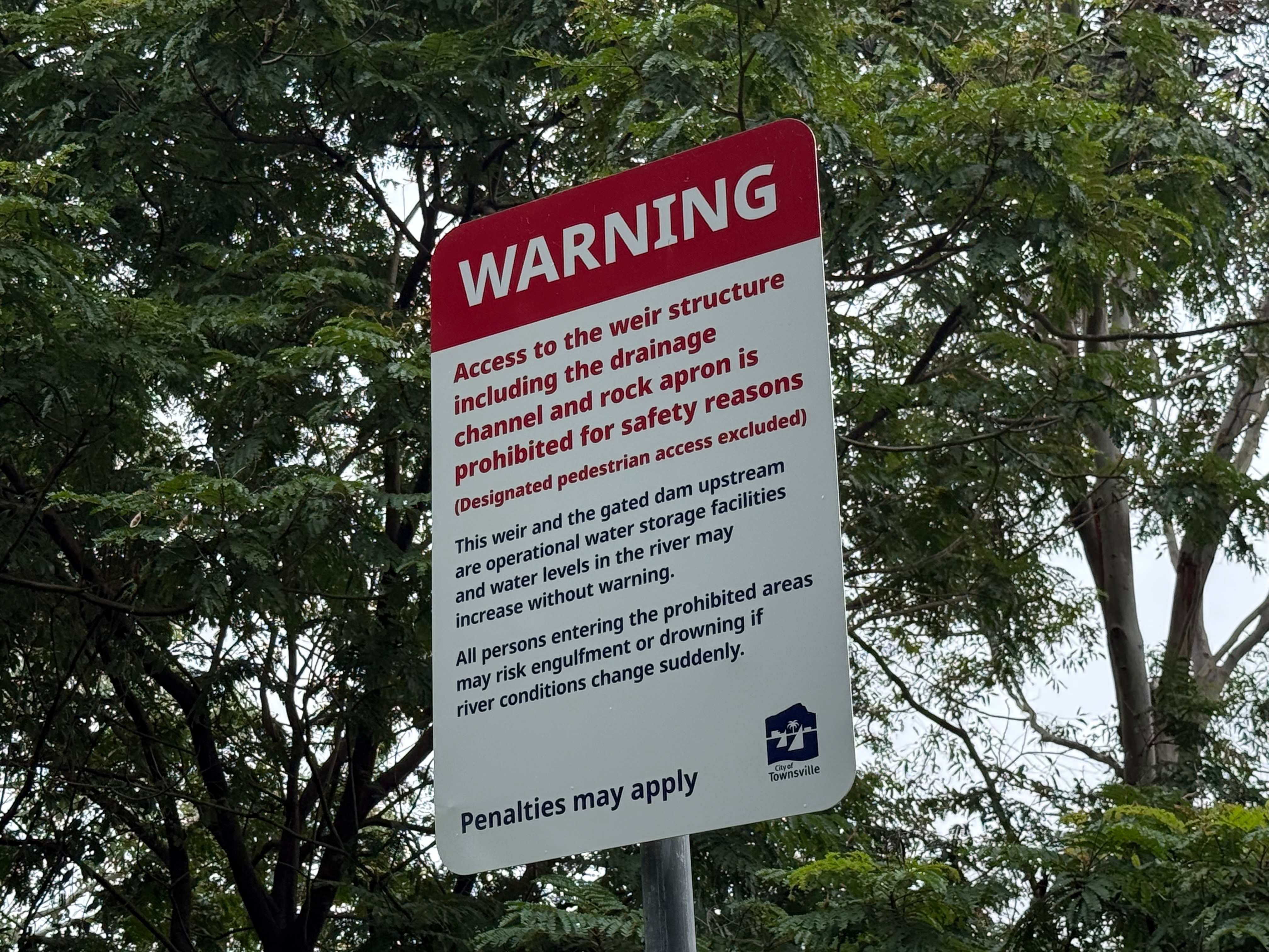 Una señal de advertencia debajo de los árboles con líneas de texto. El cartel es del Ayuntamiento de Townsville.
