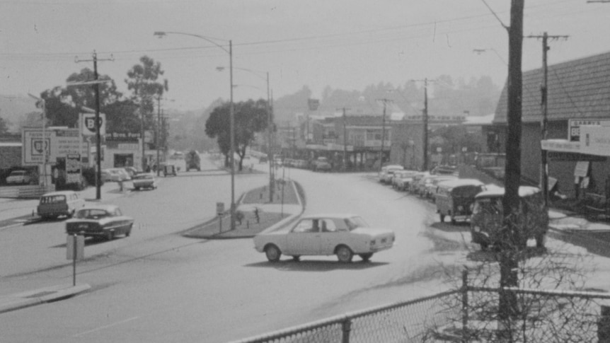 A black and white video still of a main street with cars across two lanes of seperated road