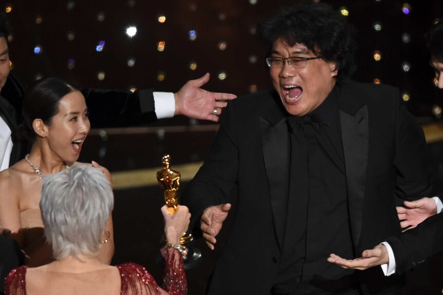 A South Korean man in a black suit smiles with his mouth open and reaches for a golden Oscars statuette.