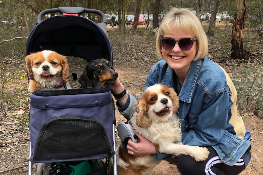 A middle-aged woman crouching on the ground holding a small dog next to a pram which contains two more small dogs.