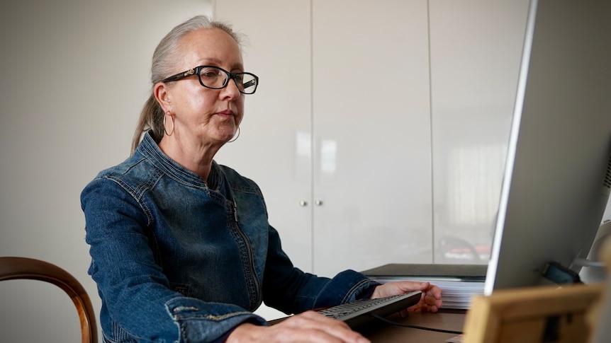 An older woman wearing a denim outfit and dark-rimmed glasses using a computer at home
