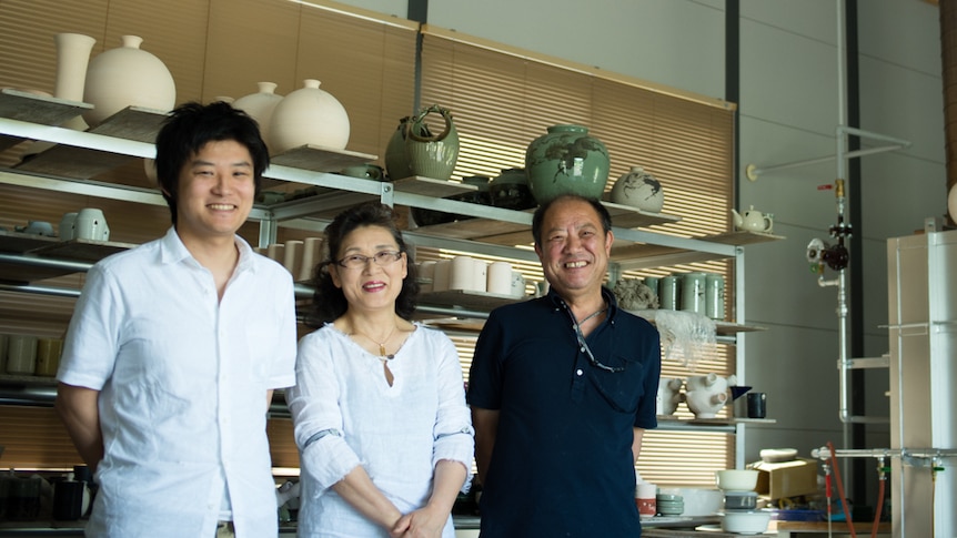 Three people smile in front of pottery on shelves.