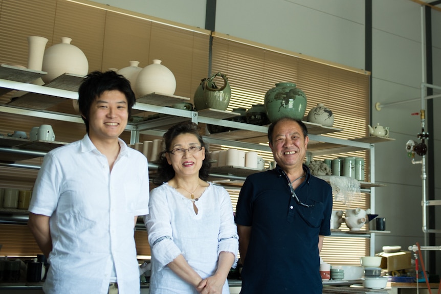 Three people smile in front of pottery on shelves.