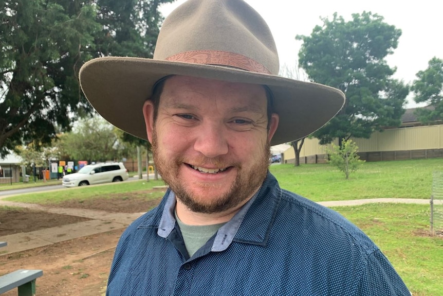 A bearded man wearing a hat and farming clothes, smiling.