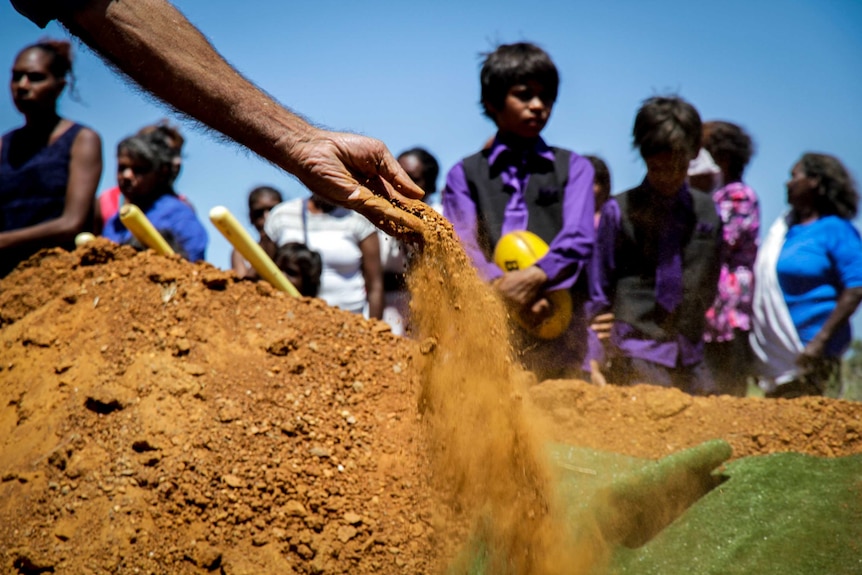 An Indigenous person throws red dirt onto a coffin at an outback funeral.