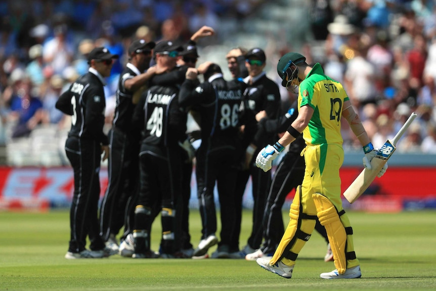 Steve Smith bows his head and carries his bat by the blade. In the background, a huddle of New Zealand players.