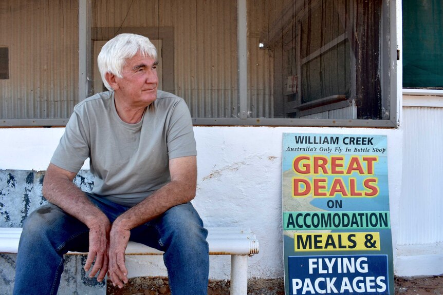 An older man with white hair sits in front of an old building.  The is taring off to his left.