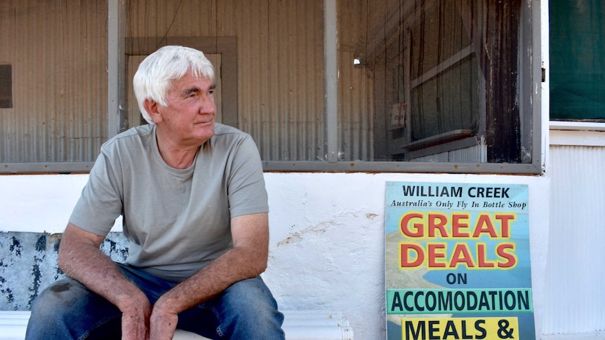 An older man with white hair sits in front of an old building.  The is taring off to his left.