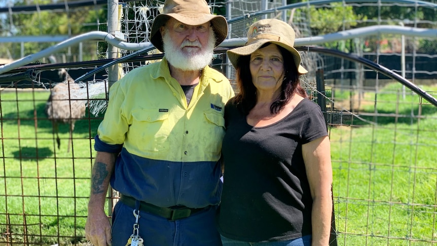 An older couple, both wearing hats, man with white bush, pose for a photo in front of enclosure, a bird in it.