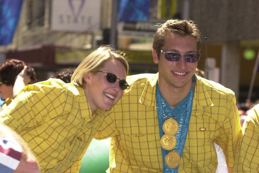 A man wears a gold jacket, sunglasses and three gold medals - a woman in a gold jacket leans in.