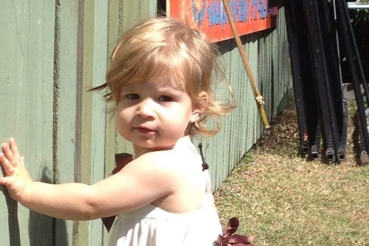 Kyanne with her arm leaning on a wooden fence, looking at the camera, brown eyes, short blonde-brown hair.