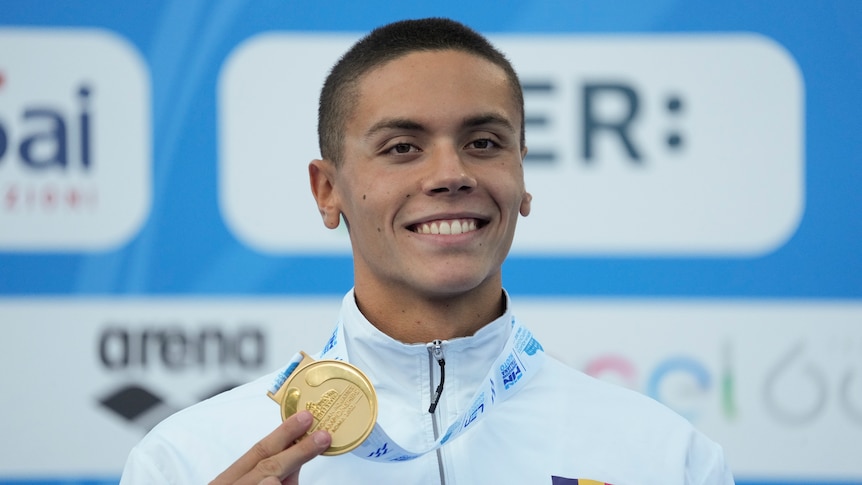 A smiling swimmer stands on the podium with a gold medal around his neck.