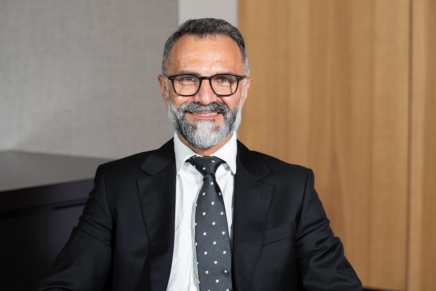 Angelo Kourtis wearing a nice suit, tie and glasses, smiling in a portrait.