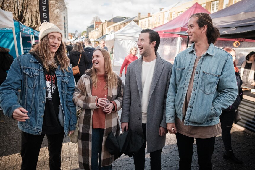 Four young people stand at an outdoor market and laugh