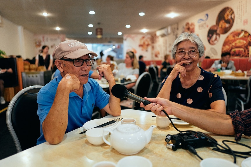 An older couple speaking into a microphone while enjoying a meal at a Chinese restaurant.