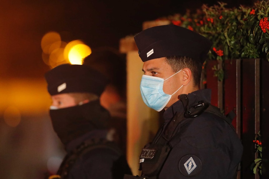 French police officers stand outside a high school in the night wearing face masks.