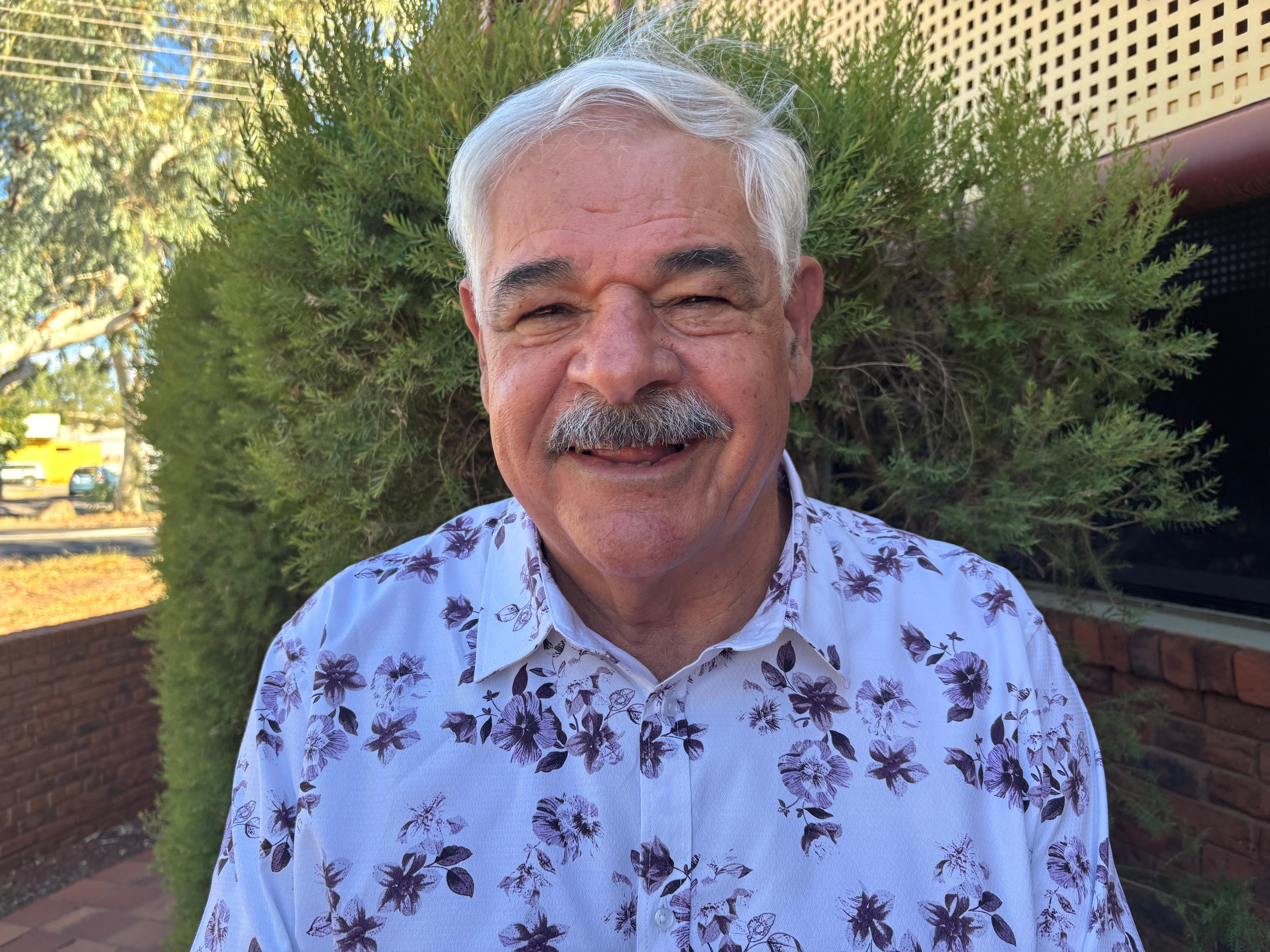 Um homem de aparência gentil, cabelos brancos e bigode fica na frente de um arbusto e sorri.