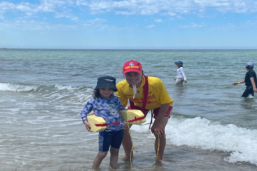 A small girl wearing bathers and a bucket hat stands next to a surf life saver in the shallows of the ocean