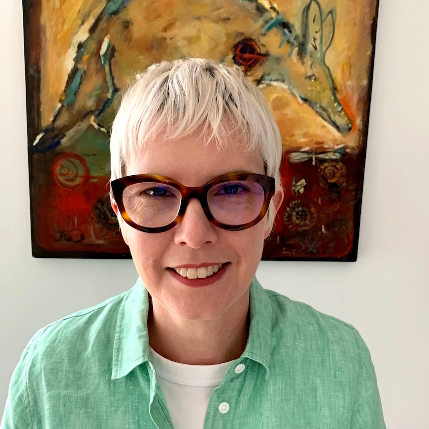 An image of Pippa Yeoman in front of a painting, wearing a green blouse and glasses.