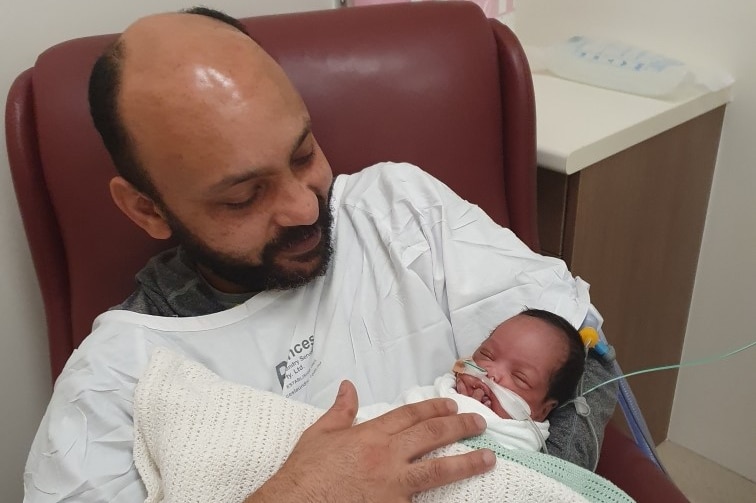 A man wearing a white hospital gown over his clothes holds a baby in his harms and smiles. There are tubes in the baby's nose.