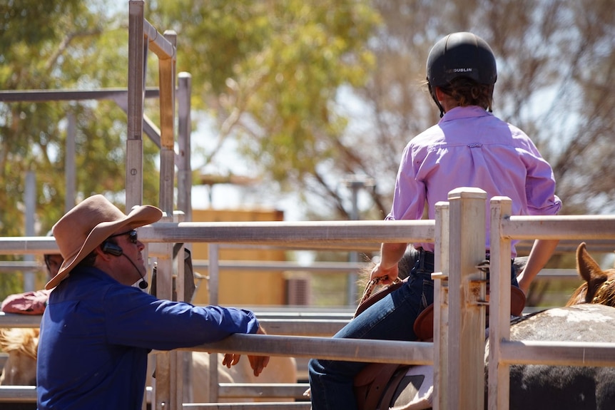 A man leans on a fence and looks at a young person on horseback.