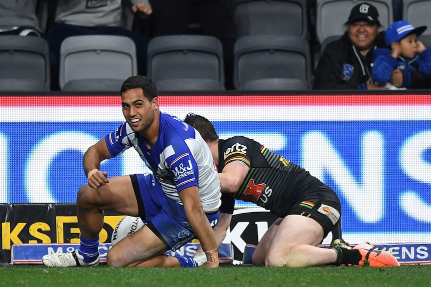 An NRL player looks up smiling after scoring a try in the corner.