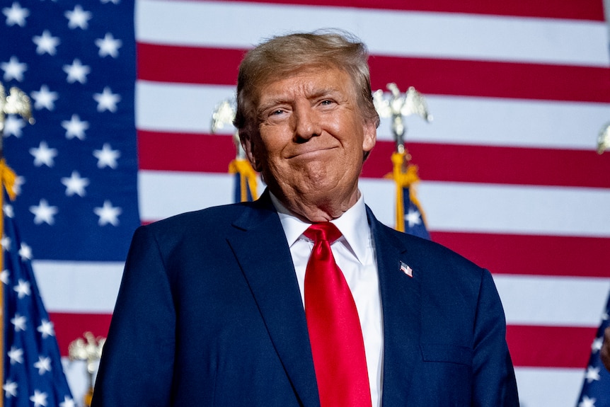 Donald Trump stands in front of a US flag background and grins.