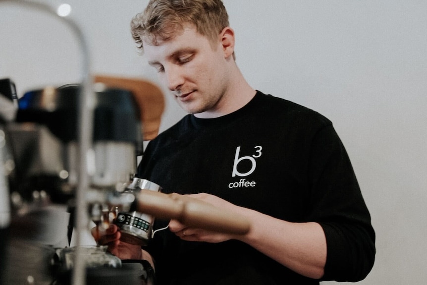 A young man wearing a black t-shirt works at a coffee machine in a cafe space.