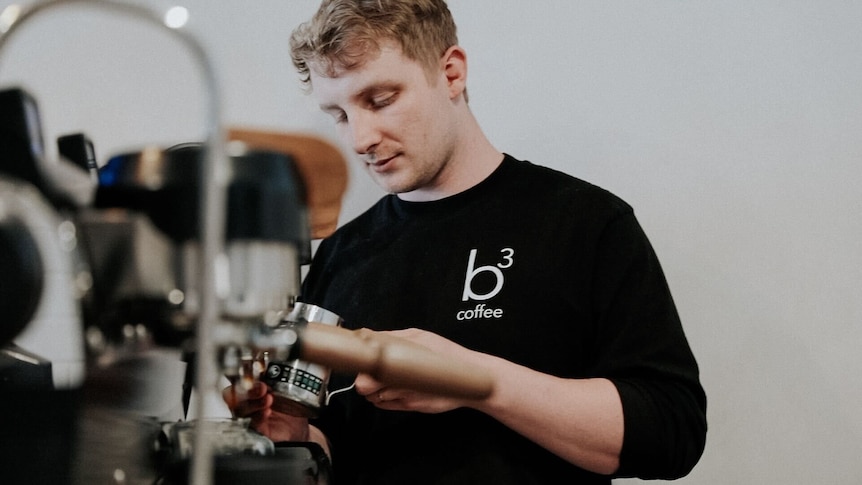 A young man wearing a black t-shirt works at a coffee machine in a cafe space.