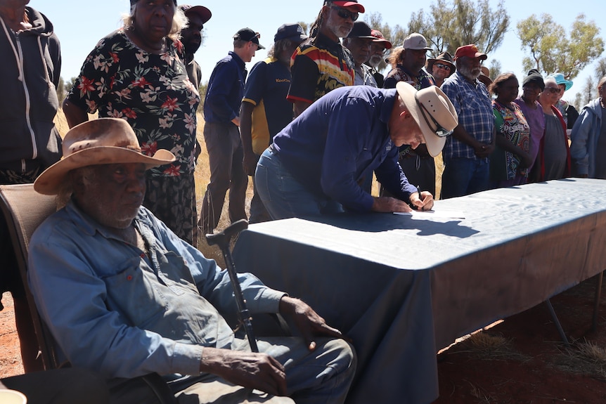 group of Indigenous people sit and stand around a long table, watching a white man in a blue shirt and akubra sign a document