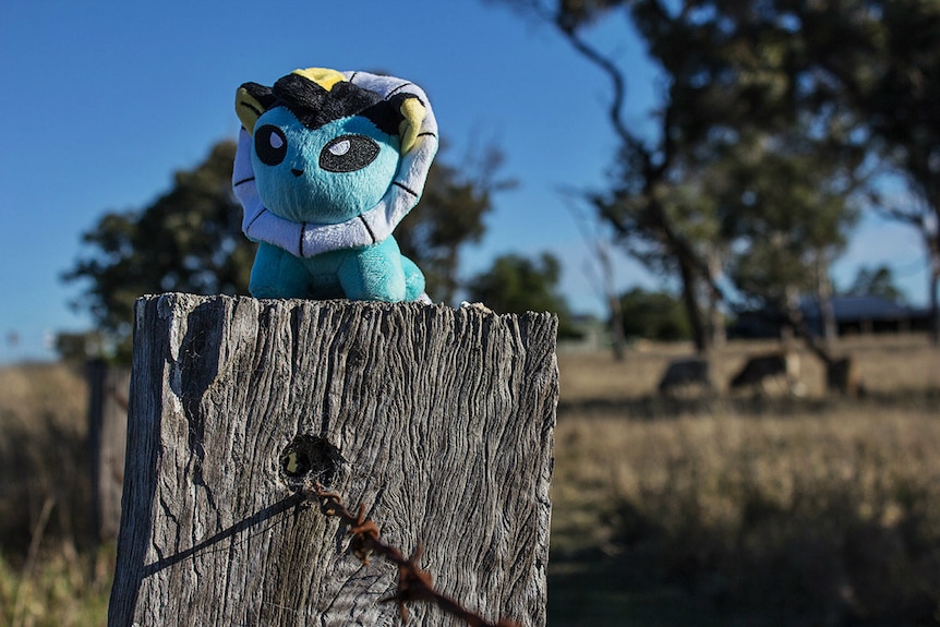 A small plush toy sits on an old wooden farm fence