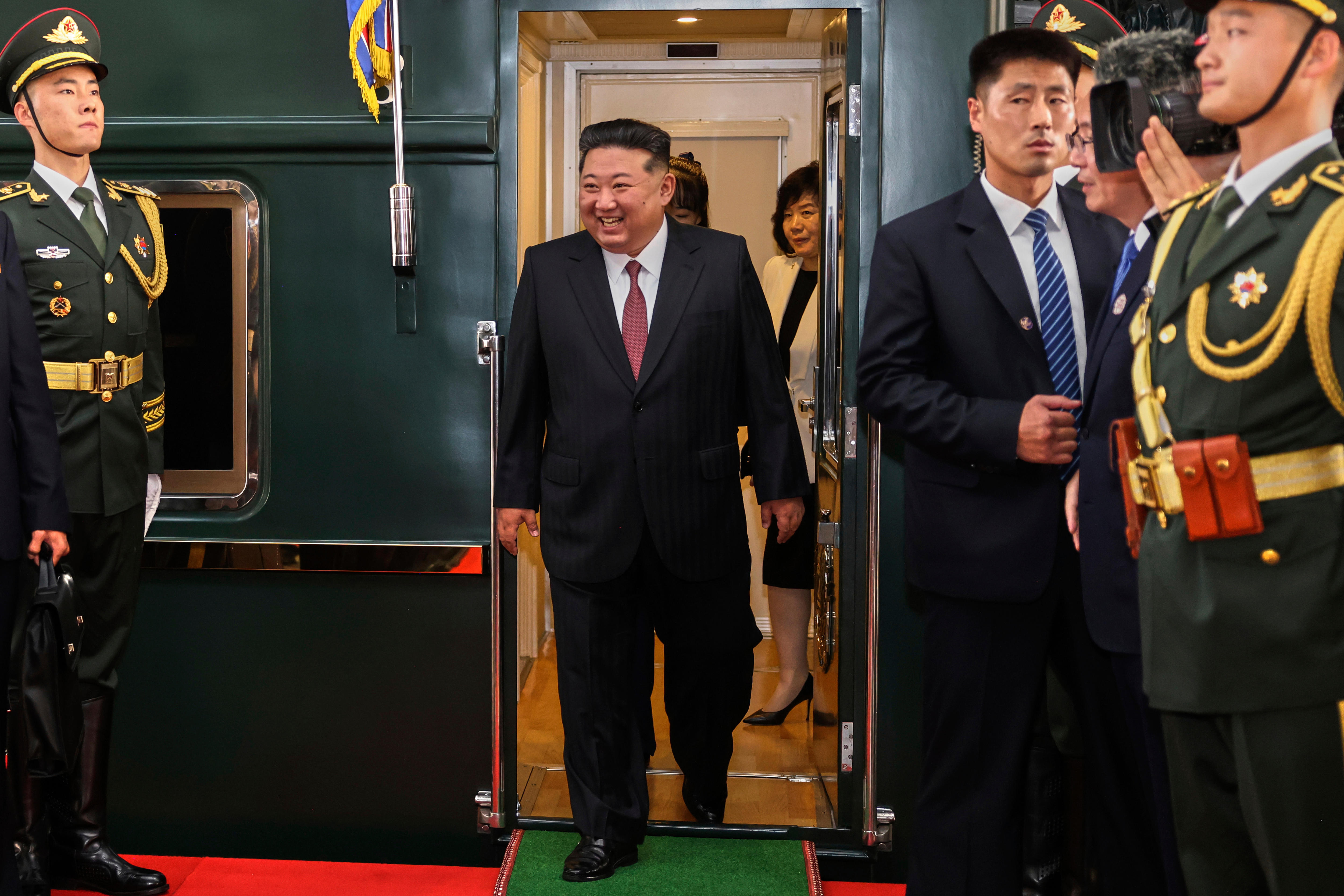 A man in a black suit steps off a green train while smiling widely, as security guards look in his direction from either side