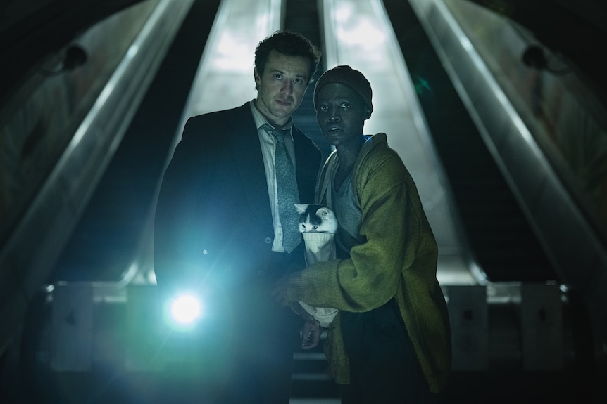 A man and a woman stand in the dark, in front of two escalators, a cat in a bag between them