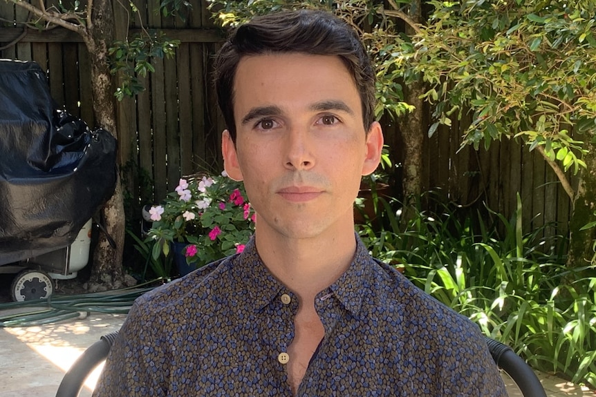 A man stares at the camera in his garden with trees behind him.