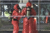 Two emegency workers in Hazmat suits stand at a large orange barrel.