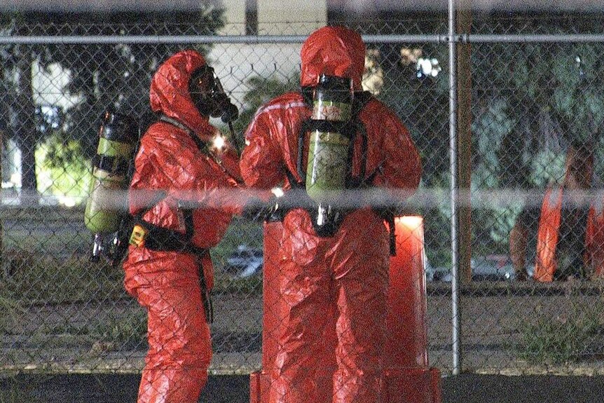 Two emegency workers in Hazmat suits stand at a large orange barrel.