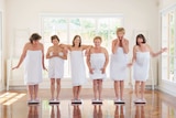 Six middle-aged women wrapped in towels pull faces while each standing on a set of scales.
