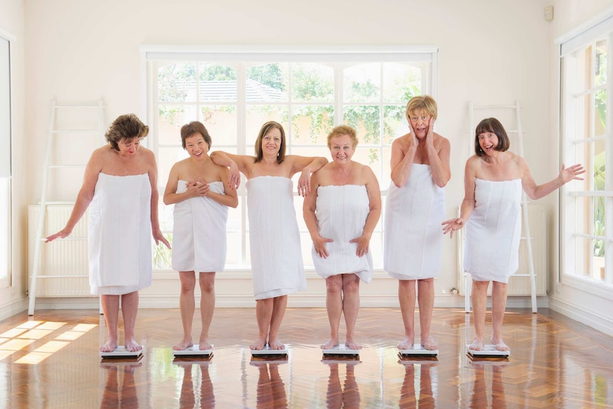 Six middle-aged women wrapped in towels pull faces while each standing on a set of scales.