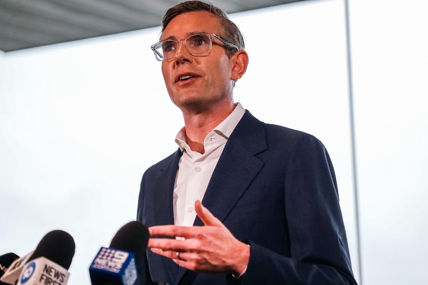 A bespectacled man with short, dark hair, wearing a dark suit – NSW Premier Dominic Perrottet – gesticulates as he speaks.
