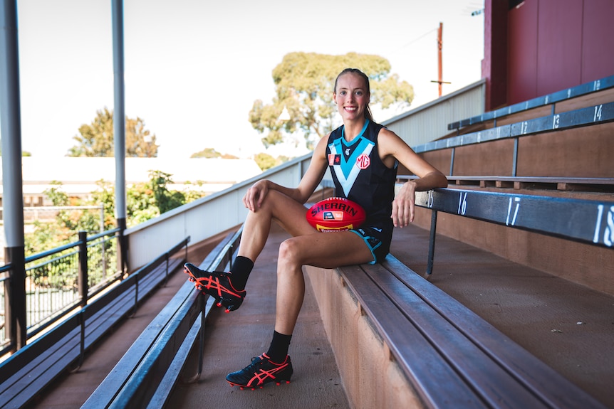 A blonde woman sitting on a chair with a red sherrin football in an AFLW uniform with black and orange boots