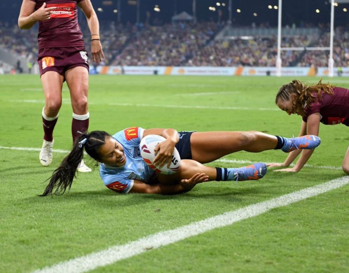 Woman in NSW blues uniform makes a try holding a football on the green field. Two females in maroon uniforms are behind her.