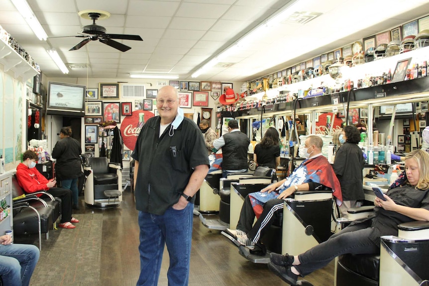 A bald man stands in a barber shop as people sit in chairs