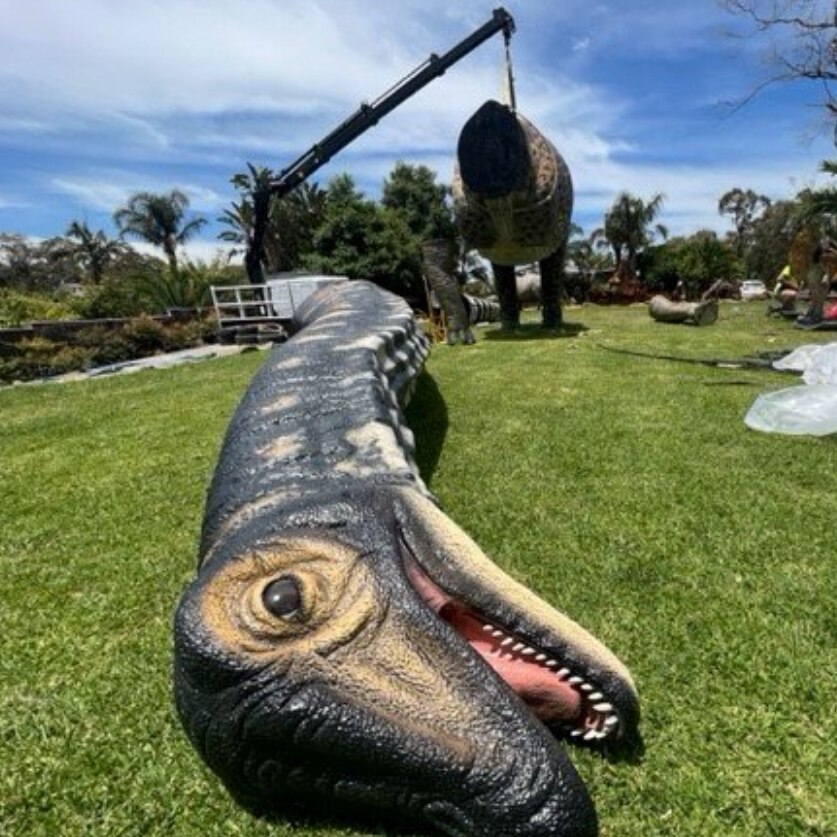 A disembodied dinosaur head lies in green grass while its neck protrudes behind 