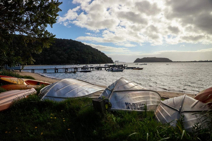 A few boats and two canoes on a patch of grass in the foreground, with Pittwater and the Great Mackerel Beach wharf behind
