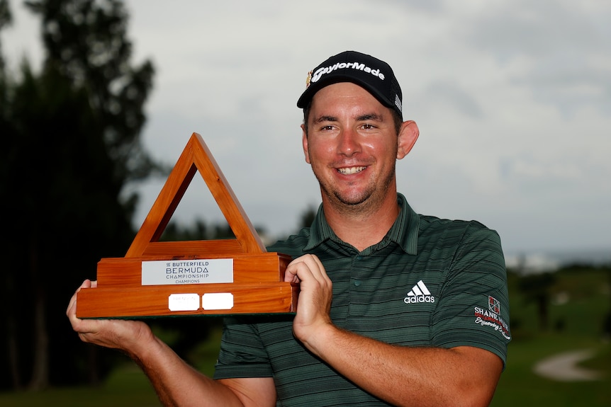 Lucas Herbert smiles while holding up the triangular Bermuda Championship trophy