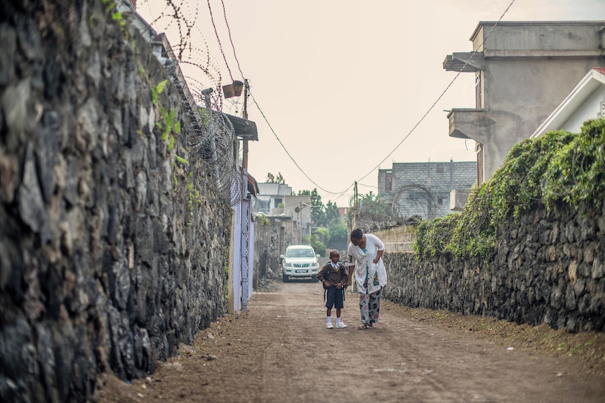 A mother walks her child, wearing a school uniform, down a dirt path towards school. 