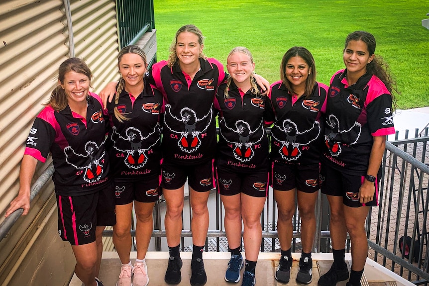 Six women hugging in pose for photo at the steps into a stadium oval.