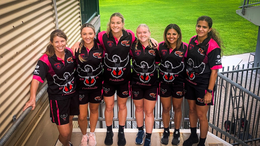 Six women hugging in pose for photo at the steps into a stadium oval.