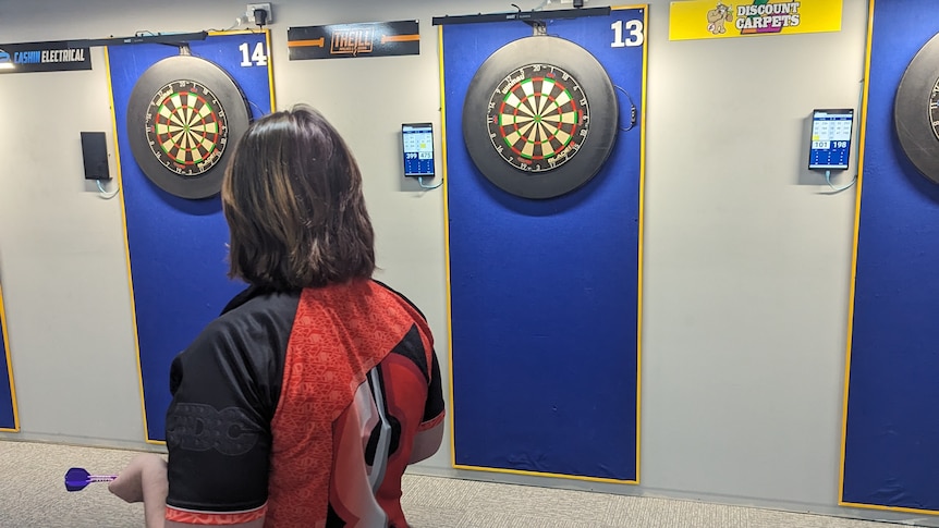 A young female darts player takes aim at the board in a club filled with darts boards